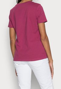 T-shirt attillato a maniche corte di un profondo colore magenta, realizzato in cotone, con una superficie liscia e un collo a giro. Pantaloni bianchi visibili.