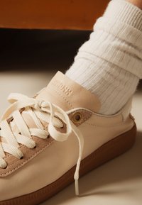 Πόδια με λευκές ριγωτές κάλτσες και μπεζ δερμάτινα αθλητικά παπούτσια με χοντρές κρεμ κορδόνια και καφέ λαστιχένιες σόλες, κοντινή άποψη.