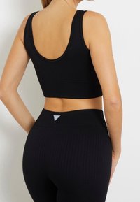 Žena ve černém žebrovaném sportovním podprsenku a odpovídajících legínách s vysokým pasem, zobrazena zezadu na pozadí bílé zdi.