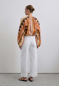 Blusa de manga larga con un vibrante patrón de hojas en naranja y negro, combinada con pantalones de pierna ancha blancos y sandalias negras de tiras.