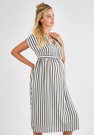 Gravid kvinde med langt blond hår iført en hvid kjole med sorte lodrette striber, der holder sin mave og rører ved sin nakke.