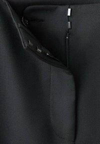 Close-up van zwarte getextureerde broek met knoop, haak- en oogsluiting en gedeeltelijk opgeloste rits op de tailleband.