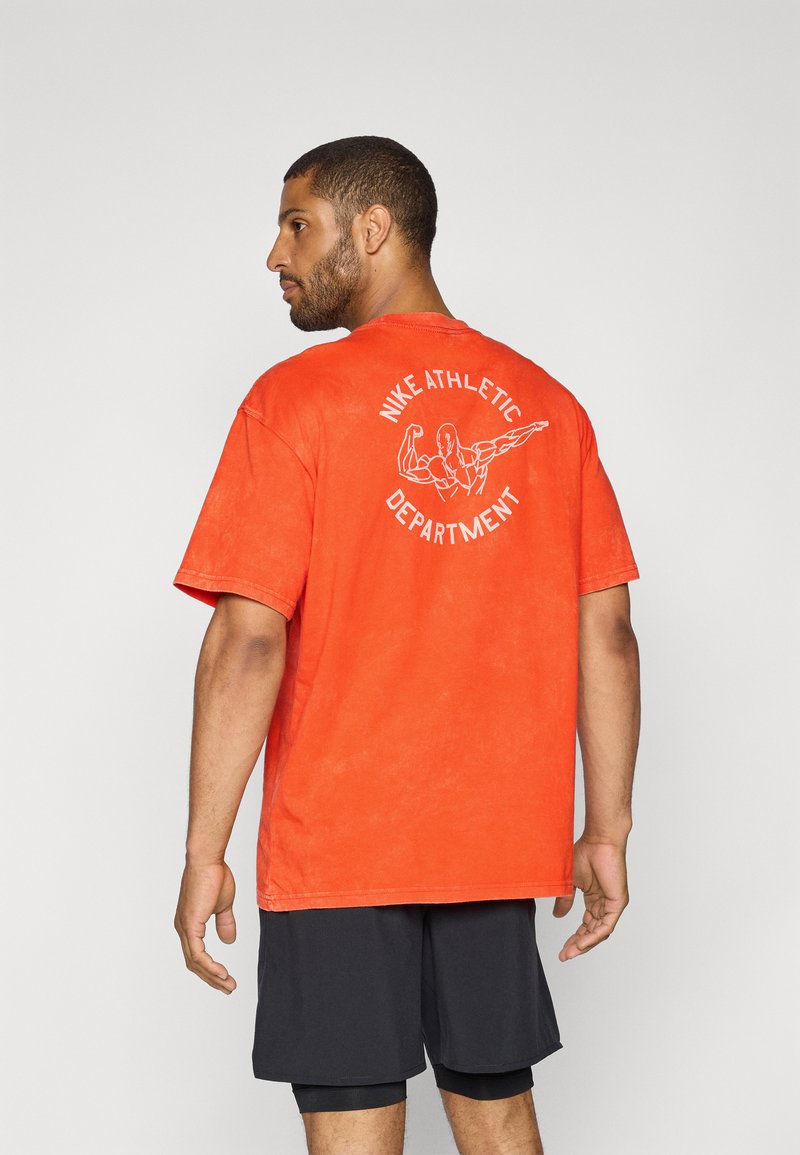Rød bomulds t-shirt med korte ærmer og en afslappet pasform. Bagsiden har hvid grafisk tekst og illustration. Bæres med sorte shorts.
