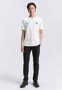 Hvid bomulds t-shirt med trykt tekst og grafik i blå og sort. Parret med sorte bukser og sorte snørebåndssko.