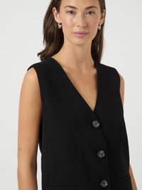 Mulher a usar um colete preto sem mangas com três botões pretos e brincos de argola prateados, contra um fundo claro e neutro.