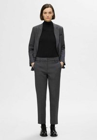 Veste de costume grise sur mesure et pantalon assorti, associés à un pull col roulé noir. Des chaussures noires complètent le look. Texture lisse, design classique.