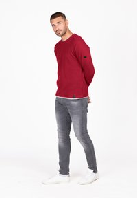 Sweatshirt en tricot rouge à côtes avec col rond et détail de tag noir, associé à un jean skinny gris déchiré et des baskets blanches.