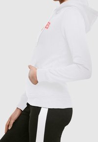 Biała bluza z kapturem, z kangurzą kieszenią, sznurkiem i czerwonym napisem. Materiał jest miękki, z ściągaczami przy mankietach i dolnej krawędzi. W zestawie z czarnymi spodniami z białym paskiem.