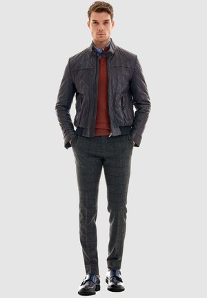 Mann trägt eine gesteppte schwarze Lederjacke, rostfarbenen Pullover, blaues Hemd, graue karierte Hose und schwarze Loafers, steht mit den Händen in den Taschen.