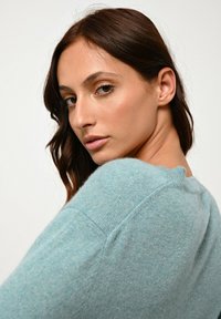 Junge Frau mit langen braunen Haaren trägt einen hellblauen Pullover und schaut über ihre Schulter vor einem schlichten weißen Hintergrund.
