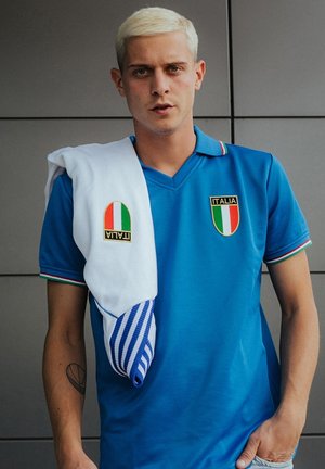 Blauwe polo met V-hals en Italiaanse patches; beschikt over groene, witte en rode accenten op de mouwen. Witte handdoek met blauwe strepen over de schouder gehangen.