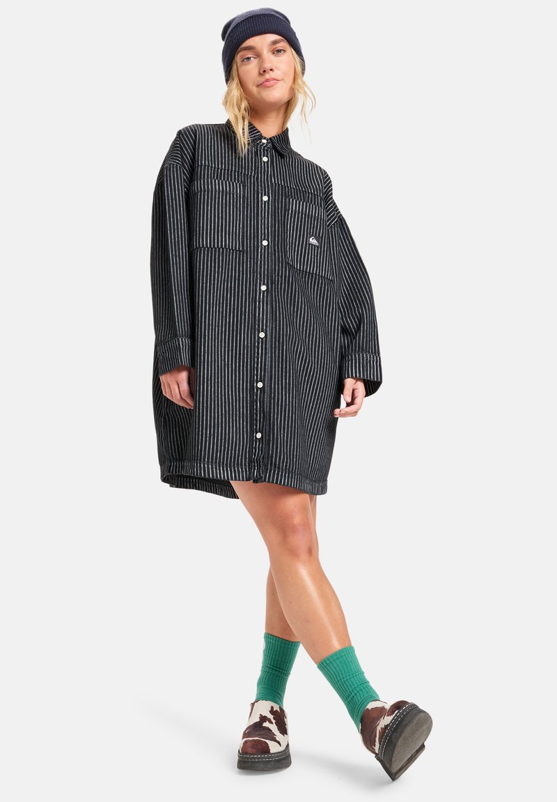Schwarz-weiße gestreifte, übergroße Hemdkleid mit Knopfverschluss, zwei Taschen auf der Vorderseite und hochgerollten Ärmeln, kombiniert mit grünen Socken und gemusterten Schuhen.