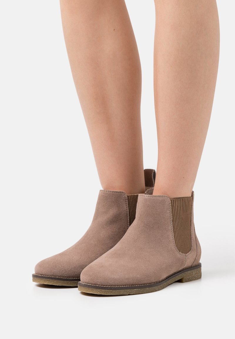 Personne portant des bottines en daim taupe avec des panneaux latéraux élastiques et des semelles plates en caoutchouc texturé, debout sur une surface claire.