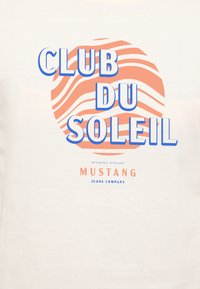 T-shirt en coton blanc présentant un texte bleu et corail avec "CLUB DU SOLEIL" et un motif ondulé en arrière-plan. Logo de la marque en bas.