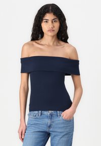 Žydros jūros spalvos off-the-shoulder bluzonė, pagaminta iš tamprios medžiagos, su plačiu iškirpimu ir prigludusia siluetu. Su deriniais šviesiai mėlynomis džinsinėmis kelnėmis.