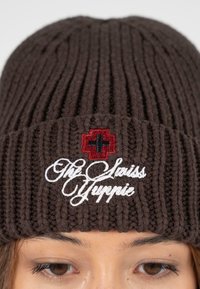 Bruine gebreide beanie met verticale ribbels, voorzien van witte cursieve tekst en een rood kruisembleem. Dikke garen zorgt voor textuur.