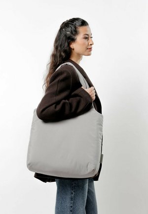 Sac fourre-tout gris en matériau doux, de forme rectangulaire, avec une large bandoulière, porté sur l'épaule sur un fond neutre.
