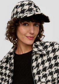 Czarnobiała czapka w wzór houndstooth wykonana z teksturowanej tkaniny, z sztywnym daszkiem. Noszona z dopasowaną kurtką w wzór houndstooth i czarnym topem.