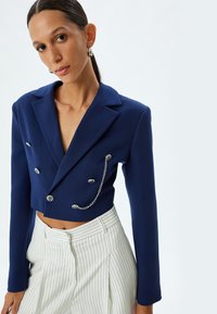 Blazer navy yang dipotong dengan leher V yang dalam, tombol ganda, dan detail rantai. Dipasangkan dengan celana tinggi bermotif garis vertikal berwarna putih.
