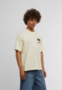 Cremefarvet bomulds T-shirt med rund halsudskæring og kortærmer. Har et sort "BEWARE" grafik på venstre brystlomme.