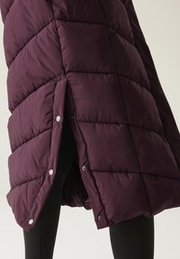 Gros plan sur une veste matelassée violet foncé avec des boutons-pression argentés partiellement ouverte sur un pantalon noir.