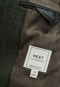 Doublure de veste marron avec une étiquette blanche Next Tailoring indiquant Regular Fit, taille 40R, fabriquée au Bangladesh, à côté d’un tissu en tweed vert.