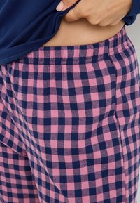 Pantalon de pyjama à carreaux rose et marine avec une taille élastique, présentant une texture douce et des poches latérales visibles.