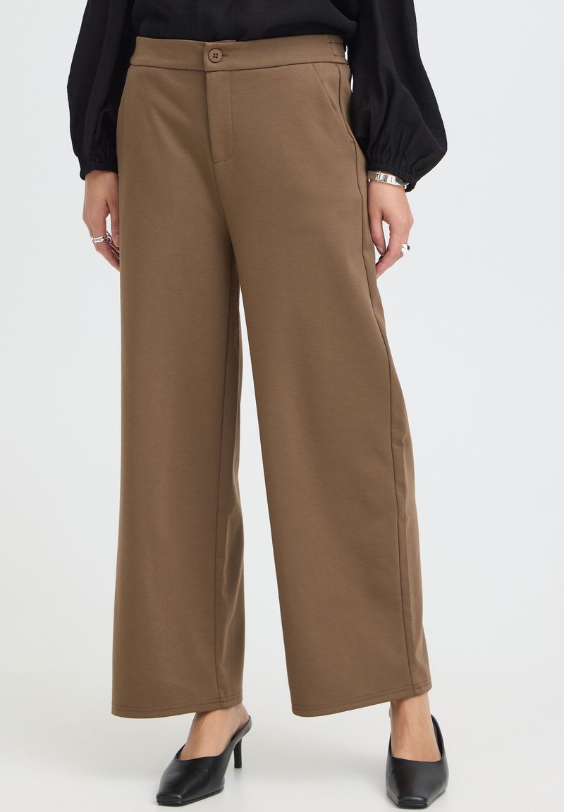 Pantalon marron à jambes larges avec bouton devant et poches, porté avec des chaussures à talons noires et un haut noir à manches longues.