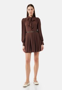 Braunes Langarmkleid mit einem Schleifenkragen, plissiertem Rock und einem schwarzen Gürtel. Weiße flache Schuhe vervollständigen das Outfit.