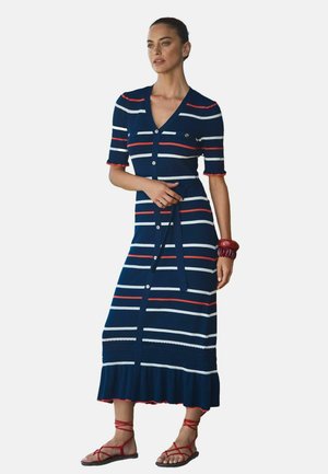 Naine, kes kannab tumesinist, punast ja valget triibulist lühikeste varrukatega nööbitavat kleiti, vööga, punaste käevõrudega ja punaste pits-up sandaalidega.