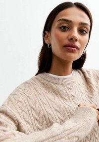 Jersey de punto beige con cuello redondo y puños acanalados. Textura suave con un aspecto de capas, usado sobre una camisa blanca.
