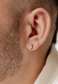 Orecchino a cerchio in argento attaccato al lobo, con una superficie liscia e lucida. Inquadratura ravvicinata della pelle texture e dei capelli corti.