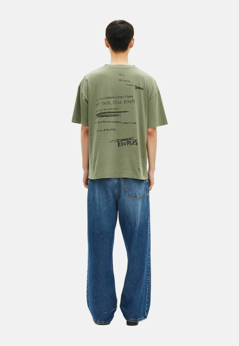 Camiseta de gran tamaño en verde oliva con texto gráfico negro en la parte posterior. Se lleva con jeans de mezclilla azules de corte holgado para un look relajado.
