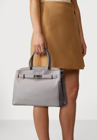 Graue Leder-Handtasche mit strukturierter Oberfläche, strukturiertem Design, doppelten oberen Griffen und einem Verschlussdetail an der Vorderseite. Kombiniert mit einem hellbraunen Rock.