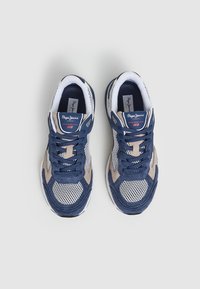 Un par de zapatillas Pepe Jeans en azul marino y beige con parte frontal de malla blanca y cordones a juego, vistas desde arriba sobre un fondo gris claro.