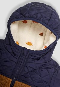 Giacca trapuntata blu navy con una sezione inferiore in pile marrone. L'interno bianco e morbido presenta motivi di dinosauri arancioni. Chiusura frontale con zip.