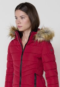 Raudona paminkštinta striukė su kailiniu gaubtu, užtrauktuku priekyje ir juodomis detalėmis. Tekstūruotos šoninės panelės suteikia dizainui papildomą charakterį.