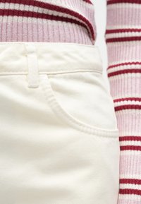 Elfenbensfarvede bomuldsbukser med en lomme foran, kombineret med en rød- og hvidstribet ribstrikket sweater. Synlige sømme og teksturerede stofdetaljer.