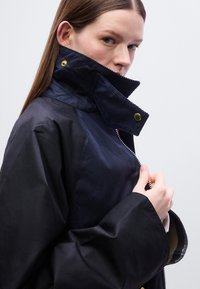 Μπουφάν σε ναυτικό μπλε με ψηλό γιακά, με φερμουάρ μπροστά και χρυσές λεπτομέρειες. Κατασκευασμένο από λείο ύφασμα με δομημένη εφαρμογή.