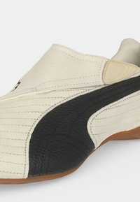 Chaussure beige profil bas avec bande texturée noire sur le côté et semelle couleur gomme, avec des détails cousus et un rabat supérieur fixé.