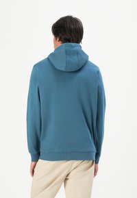 Uomo che indossa una felpa con cappuccio color teal e pantaloni beige, in piedi con le spalle rivolte verso uno sfondo bianco puro.