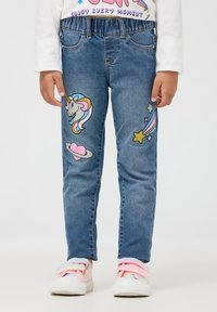 Dżinsy z elastycznym paskiem w kolorze niebieskim, z naszywkami w kształcie jednorożca, serca i gwiazdy w pastelowych kolorach z przodu. Białe trampki z pastelowymi paskami.