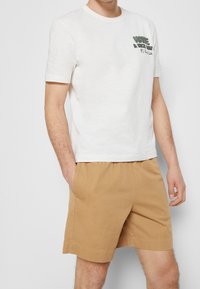 Camiseta blanca de manga corta con texto gráfico verde en el frente, combinada con pantalones cortos elásticos color marrón claro. Mezcla de algodón, ajuste relajado.