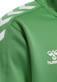 Zielony rękaw sportowej kurtki z białymi pasami w kształcie szewroń i częścią białego logo Hummel na piersi.