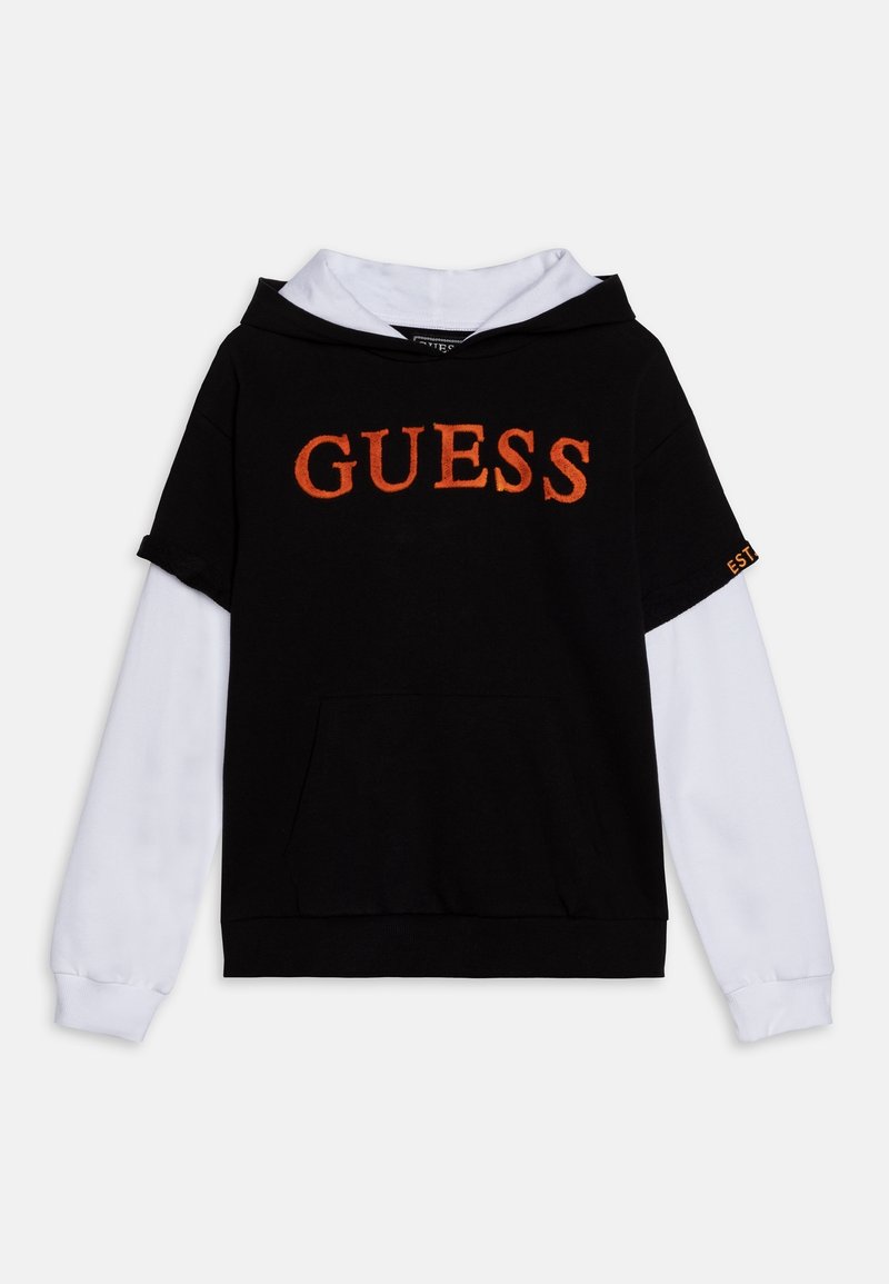 Sweat à capuche noir à manches courtes superposé sur une chemise blanche à manches longues, avec le mot "GUESS" en orange sur le devant.