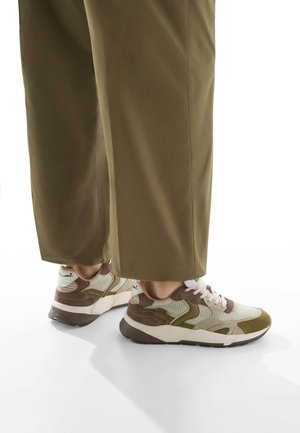 Sneaker mit einem Obermaterial aus Mesh in hellem Creme und olivgrün, braunen Akzenten und einer dicken weißen Sohle, kombiniert mit lockeren olivfarbenen Hosen.