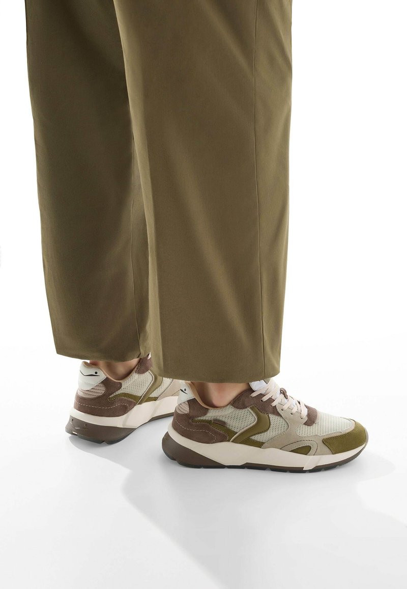 Sneakers con tomaia in rete color crema chiaro e verde oliva, dettagli marroni e suola bianca spessa, abbinati a pantaloni larghi verde oliva.
