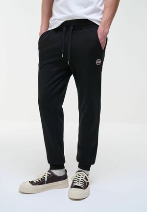 Uomo che indossa pantaloni da jogging neri con coulisse e sneakers bianche e nere, in piedi con le mani nelle tasche su sfondo bianco.