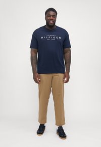 Tumši zilas kokvilnas T-krekls ar "TOMMY HILFIGER" logotipu, apvienots ar bēšām biksēm un melniem apaviem. Modelis stāv frontālajā pozīcijā.