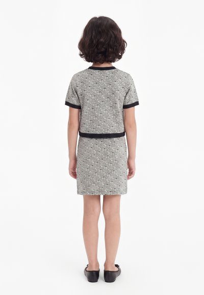 Copil cu păr scurt și creț stă cu fața spre spate, purtând un top gri cu mânecă scurtă asortat și o fustă cu margini negre și balerini negri.